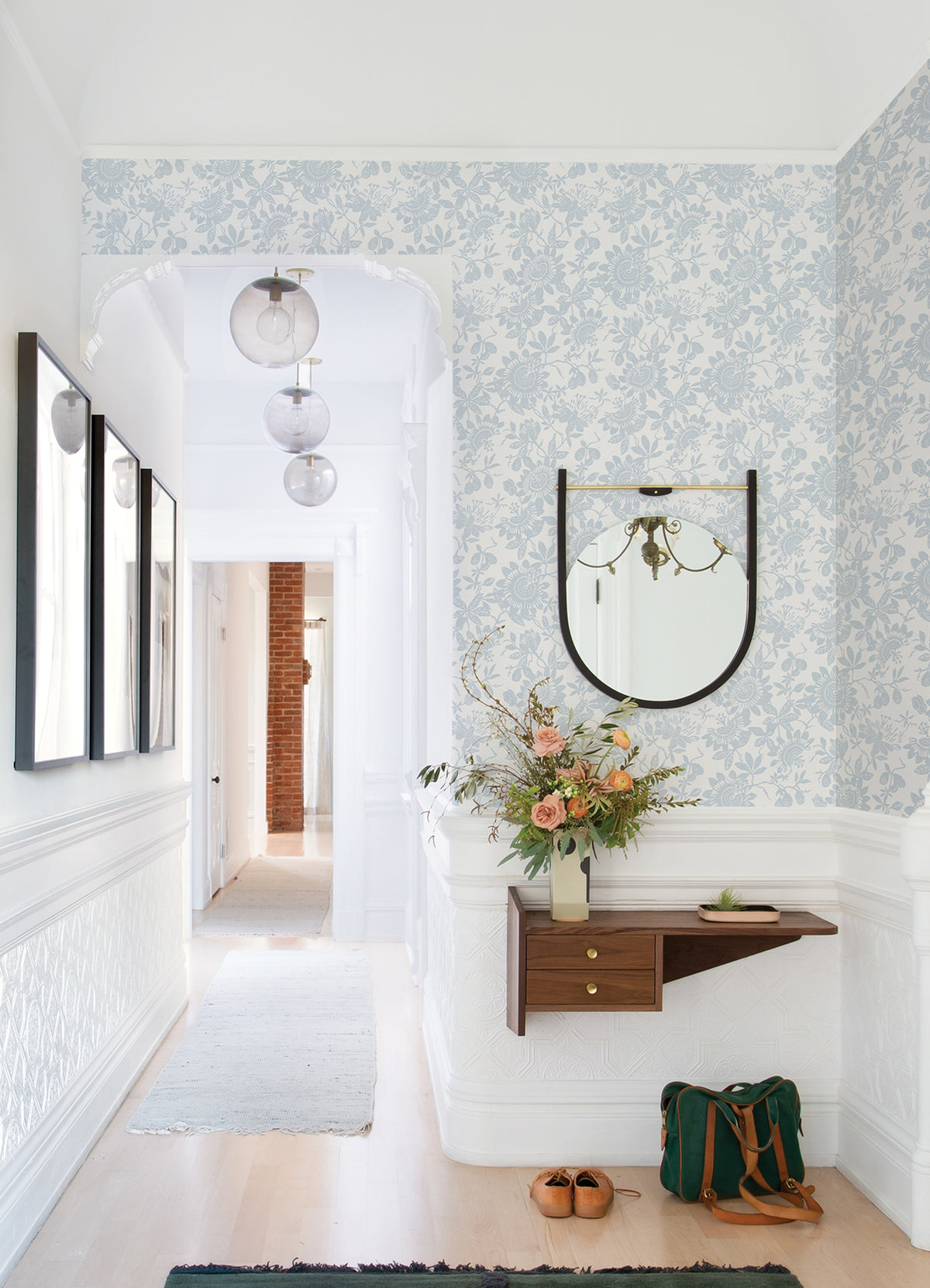 A bright hallway featuring Helen Floral Trail Wallpaper by York Wallcoverings, a round mirror, wall art, a floating shelf displaying a vase with flowers and a book, brown shoes, and a green bag resting on the light wood floor. Three glass pendant lights hang from the ceiling.