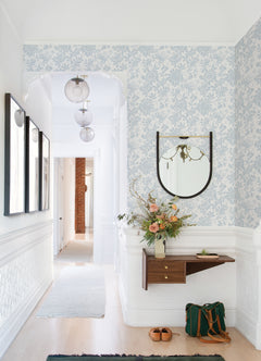 A bright hallway featuring Helen Floral Trail Wallpaper by York Wallcoverings, a round mirror, wall art, a floating shelf displaying a vase with flowers and a book, brown shoes, and a green bag resting on the light wood floor. Three glass pendant lights hang from the ceiling.