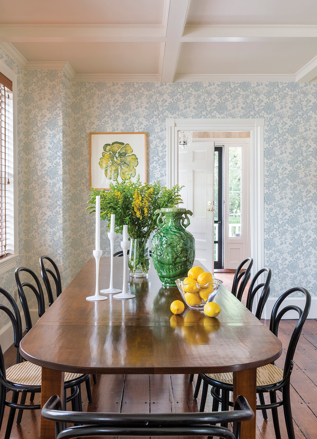 A dining room featuring a wooden table, black chairs, white candles, a green vase filled with yellow flowers, and a bowl of lemons. The background showcases York Wallcoverings’ Helen Floral Trail Wallpaper in light blue floral pattern, along with an open door.