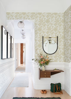 A bright hallway features Helen Floral Trail Wallpaper by York Wallcoverings, a round mirror above a floating shelf adorned with flowers and books, wall art, and a green bag with brown shoes resting on the light wood floor.