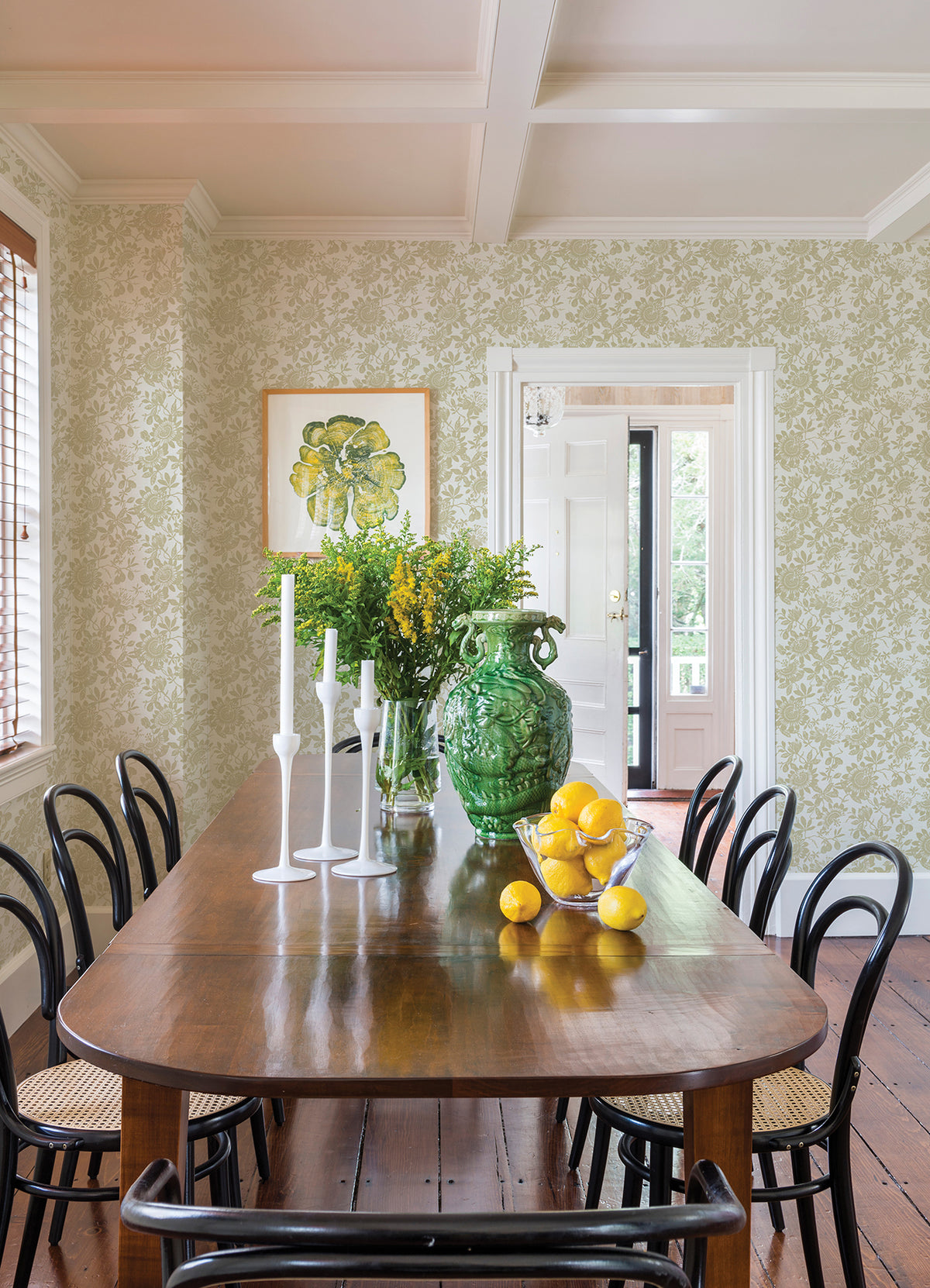 A wooden dining table with black chairs, white candlesticks, a green vase filled with yellow flowers, and a bowl of lemons is set in a bright room featuring Helen Floral Trail Wallpaper by York Wallcoverings and framed art.