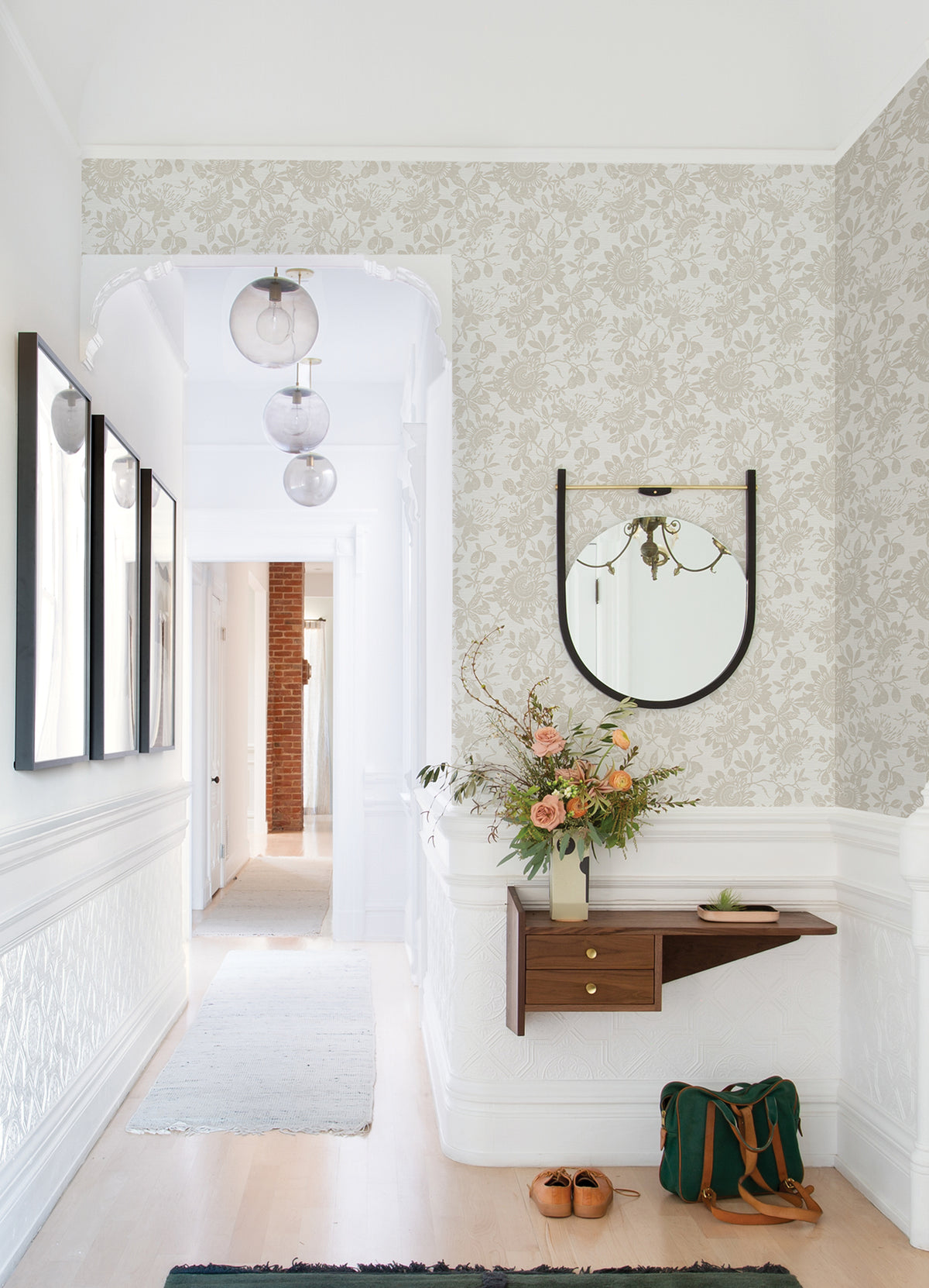 Bright hallway featuring Helen Floral Trail Wallpaper by York Wallcoverings, a wall mirror, and a small wooden shelf holding a vase of flowers; shoes and a green bag rest on the light wood floor, while three globe pendant lights hang from the ceiling.