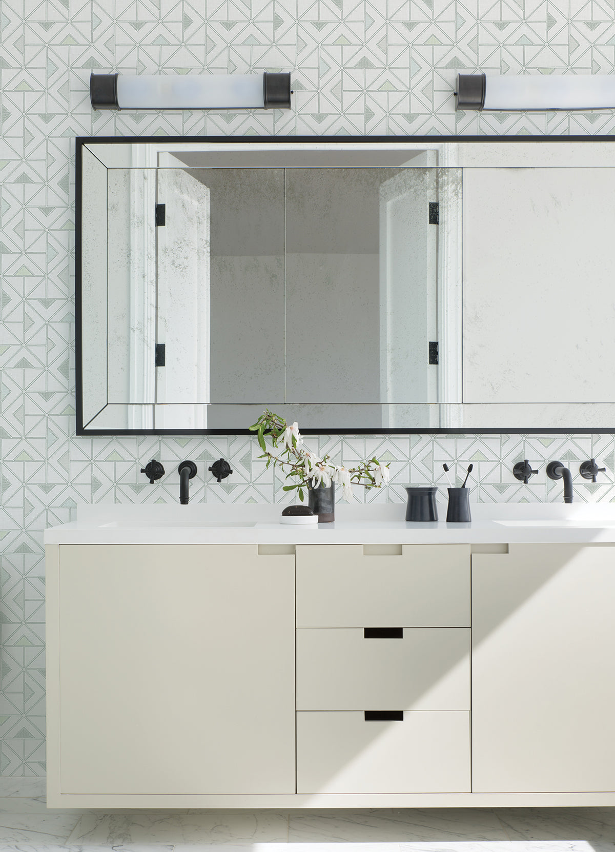 Modern bathroom featuring a large mirror, Jekyl Triangles Wallpaper by York Wallcoverings on the walls, a white vanity with black hardware, two wall lights above, and minimal accessories like a vase with green branches.