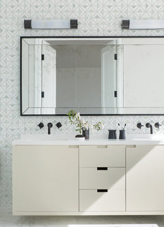 Modern bathroom featuring a large mirror, Jekyl Triangles Wallpaper by York Wallcoverings on the walls, a white vanity with black hardware, two wall lights above, and minimal accessories like a vase with green branches.