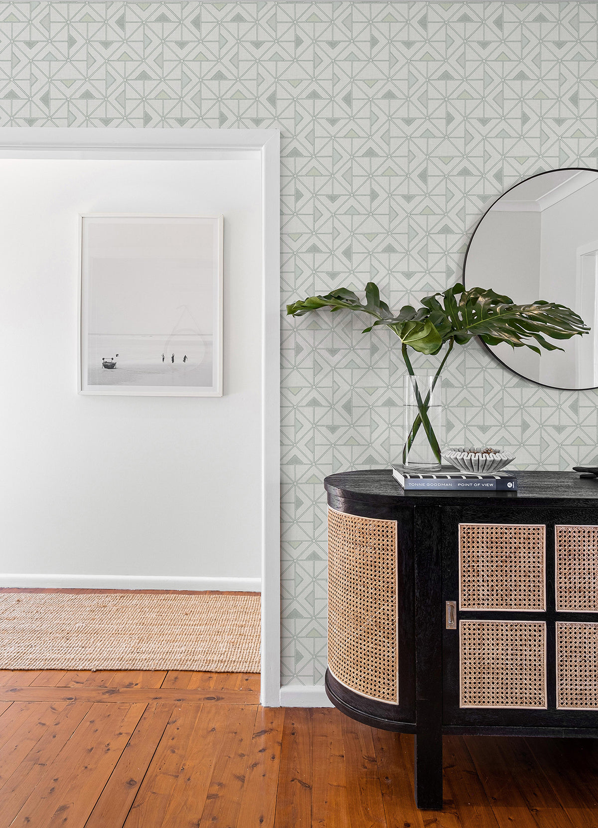 A modern interior featuring York Wallcoverings’ Jekyl Triangles Wallpaper, complemented by a round mirror, a black cabinet with cane doors, a plant in a vase, and a framed photo on the white wall. The wooden floor leads into the next room with a woven rug.