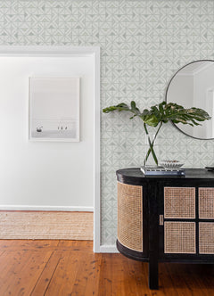 A modern interior featuring York Wallcoverings’ Jekyl Triangles Wallpaper, complemented by a round mirror, a black cabinet with cane doors, a plant in a vase, and a framed photo on the white wall. The wooden floor leads into the next room with a woven rug.