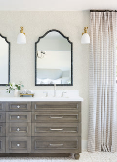 Bright bathroom featuring Scout Floral Ogee Wallpaper by York Wallcoverings, double mirrors above a wood vanity with drawers, two white light sconces, a vase with flowers, and floor-length striped curtains beside the sink.