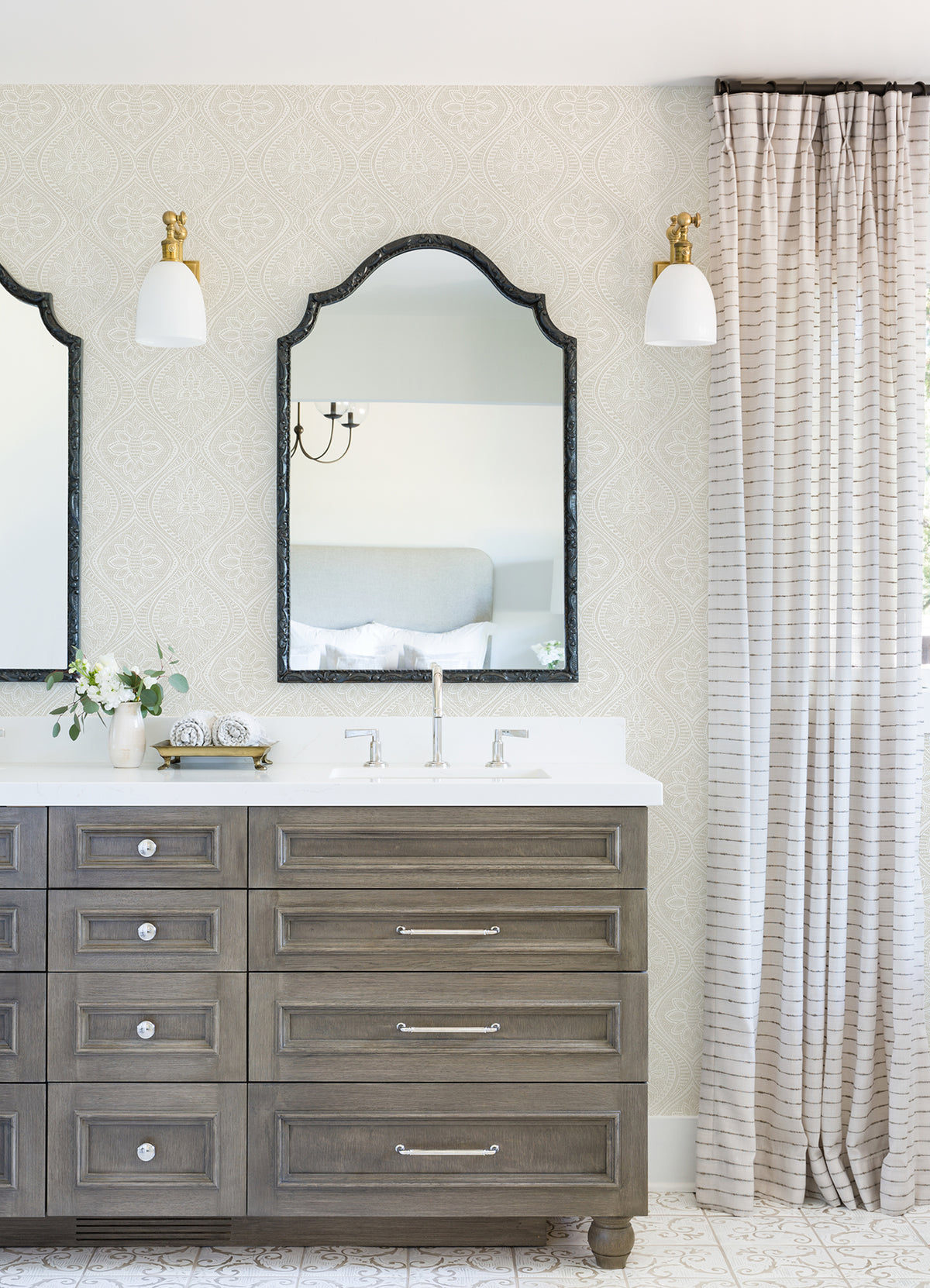 A bathroom featuring a double sink vanity, two mirrors, brass light fixtures, York Wallcoverings’ Scout Floral Ogee Wallpaper in a neutral palette, a small vase with flowers, and tall striped curtains on the right side.