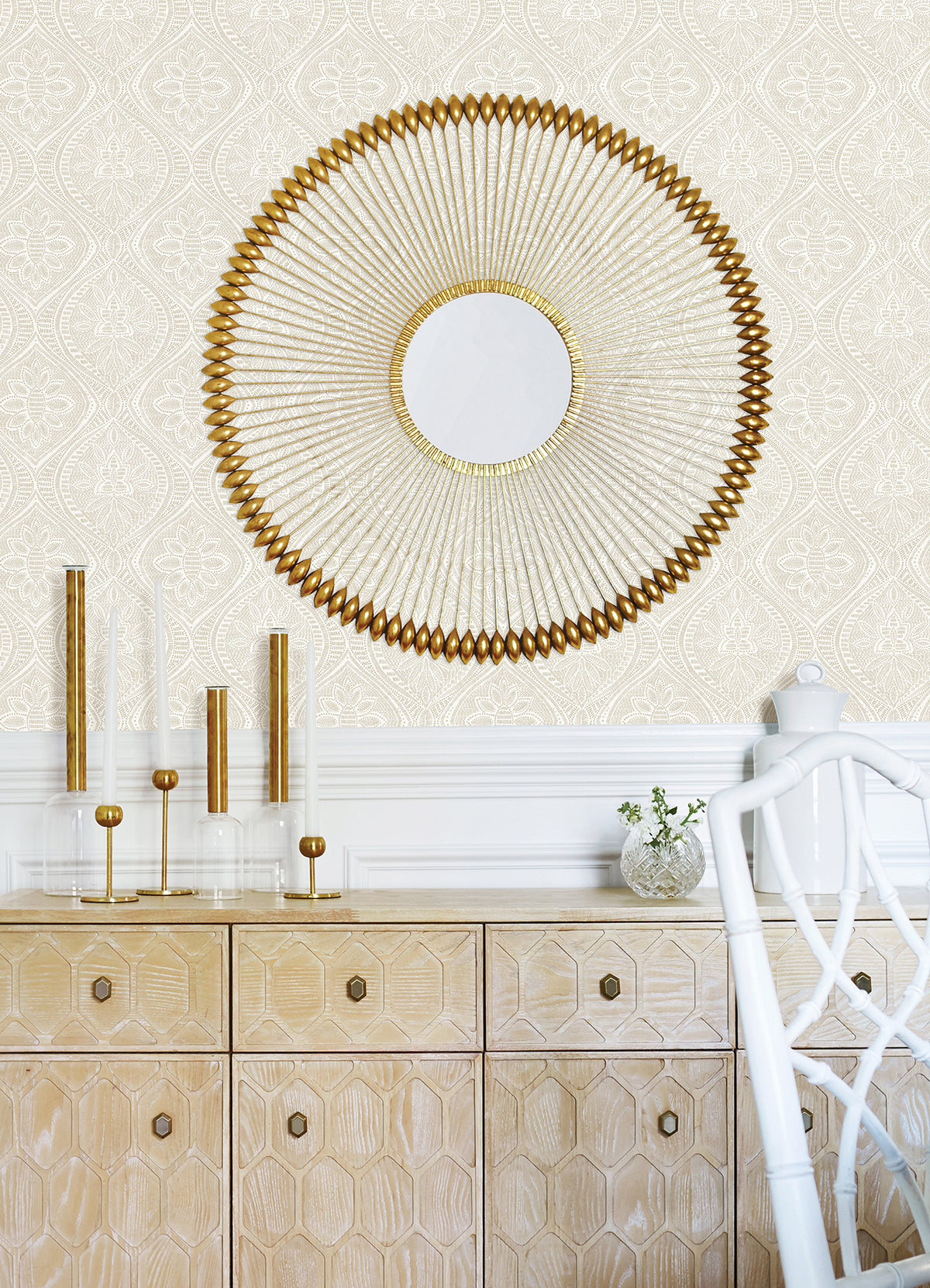 A feature wall adorned with Scout Floral Ogee Wallpaper by York Wallcoverings forms a stylish backdrop above a light wood cabinet. On the cabinet are modern gold and glass candlesticks, a small vase with greenery, and a white chair.