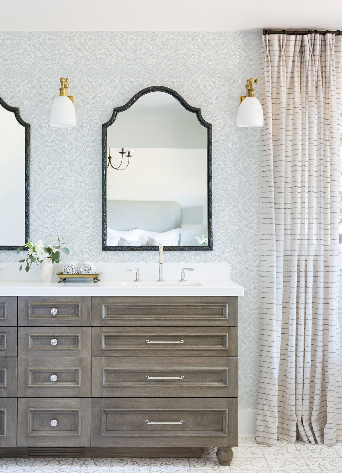 Elegant bathroom with a double vanity, two mirrors, and wall sconces. The space is finished with York Wallcoverings’ Scout Floral Ogee Wallpaper, neutral curtains, and fresh flowers on the countertop for a serene and sophisticated look.