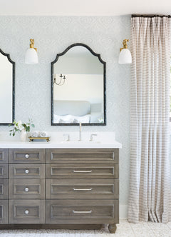 Elegant bathroom with a double vanity, two mirrors, and wall sconces. The space is finished with York Wallcoverings’ Scout Floral Ogee Wallpaper, neutral curtains, and fresh flowers on the countertop for a serene and sophisticated look.