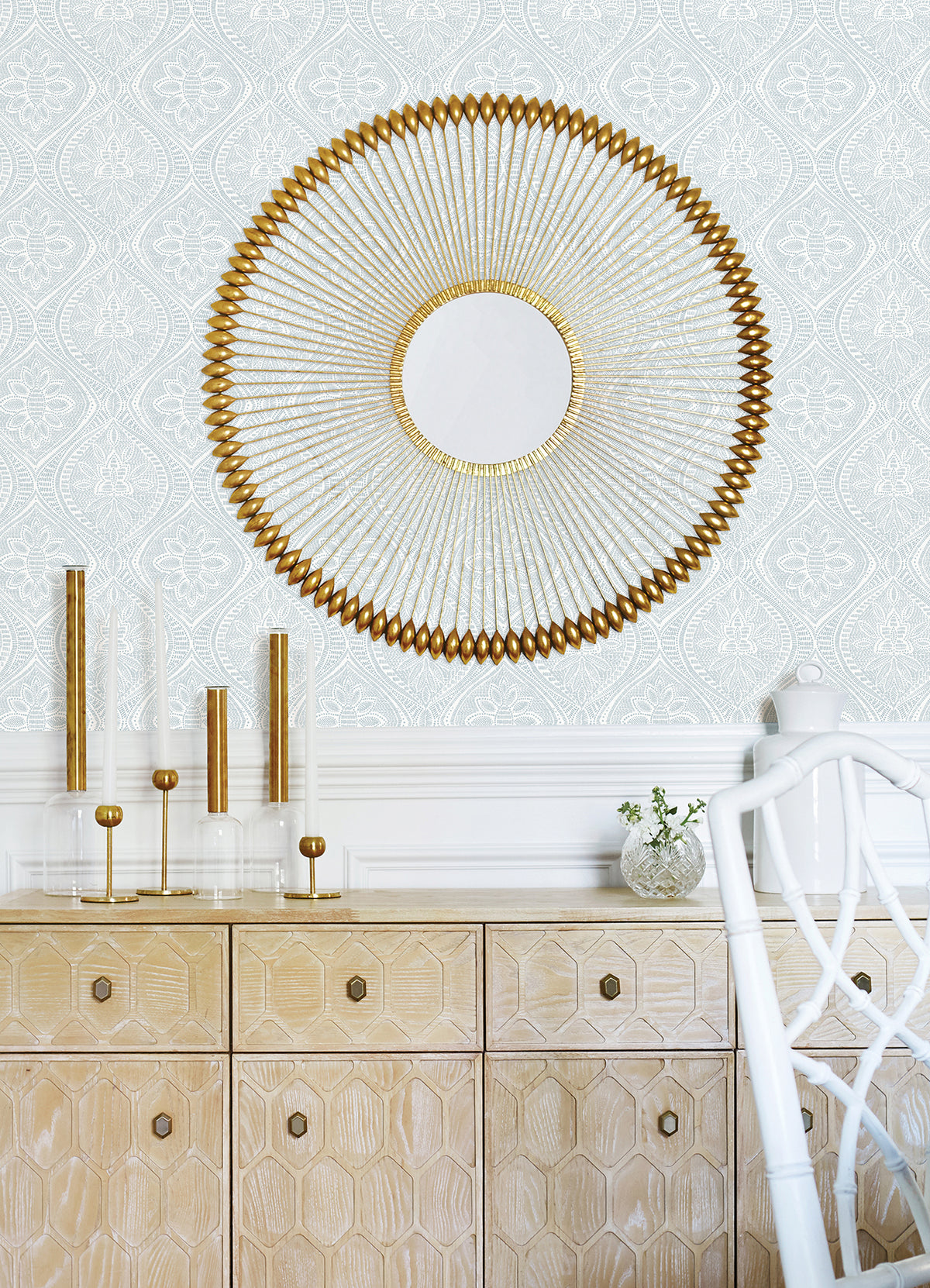 A wall covered in Scout Floral Ogee Wallpaper by York Wallcoverings is the backdrop for a light wood cabinet with a geometric pattern. Above the cabinet hangs a round gold decorative mirror, while on top of the cabinet sit gold and glass candle holders and a small vase with greenery. A white chair is partially visible on the right.
