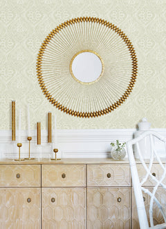 A gold sunburst mirror hangs above a light wood cabinet with geometric patterns, which displays gold and glass candle holders and a vase filled with greenery. In the foreground sits a white chair, while the walls are adorned with Scout Floral Ogee Wallpaper by York Wallcoverings in the background.