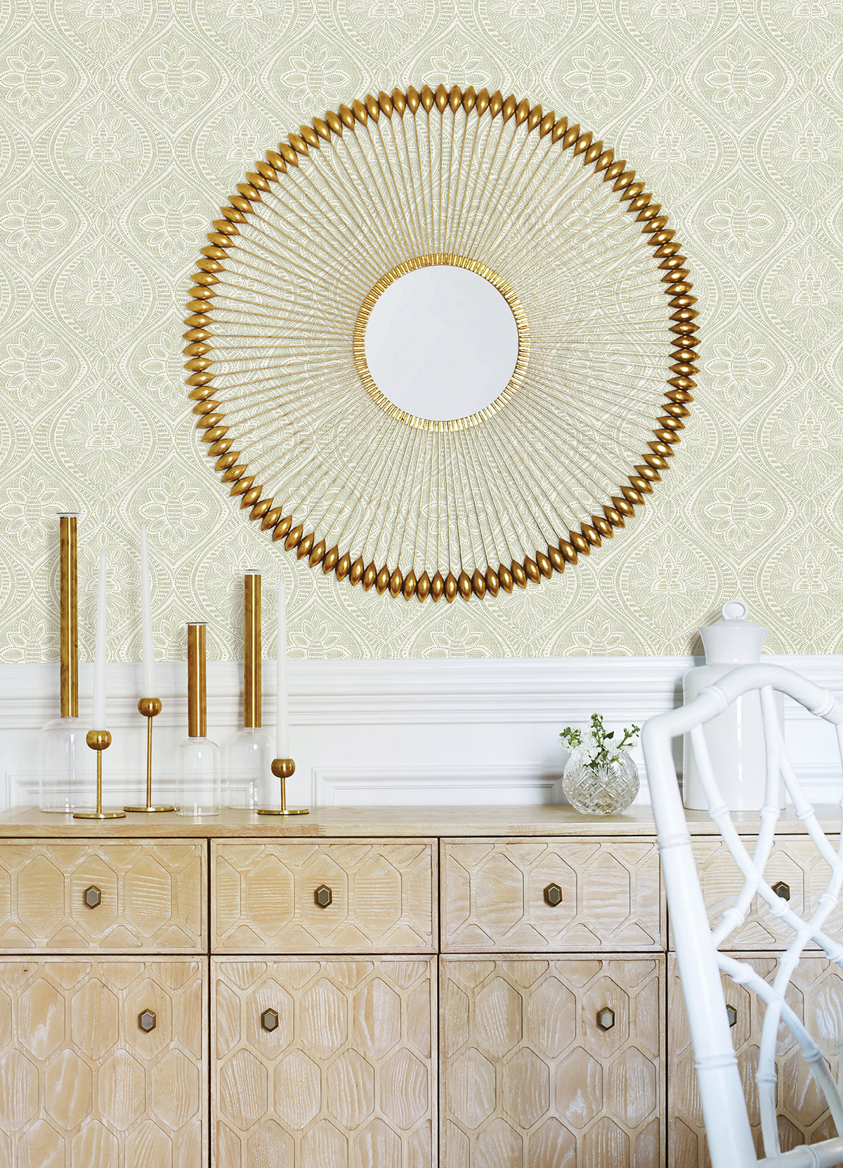 A round gold decorative mirror hangs on a wall adorned with Scout Floral Ogee Wallpaper by York Wallcoverings above a light wood cabinet. The cabinet displays gold-accented candleholders and a small vase with greenery, while a white chair is partially visible.
