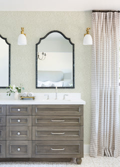 A bathroom vanity with wood drawers, double faucets, two mirrors, wall sconces, a small vase with flowers, a clock, and patterned curtains is set against Scout Floral Ogee Wallpaper by York Wallcoverings.