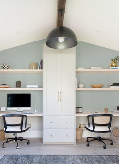 Transformed Sentence: Modern home office featuring two desks, black office chairs, a large overhead light fixture, built-in white shelves displaying books and decor, a desktop computer, and neutral minimalist accents—all brought together by Glynn Chevron Wallpaper from York Wallcoverings.