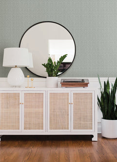 A modern sideboard sits against a wall adorned with Glynn Chevron Wallpaper by York Wallcoverings, topped with a white lamp, two gold candlesticks, potted plants, and a record player; above hangs a large round mirror, while a tall plant stands beside it on the wooden floor.