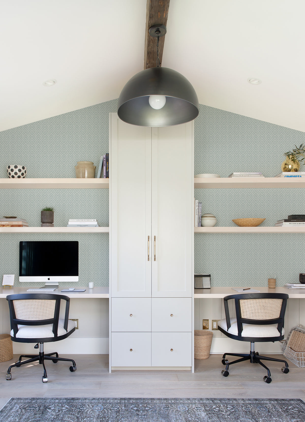 Modern home office with two chairs at a shared white desk, built-in shelves with decor, a computer, and storage in the center. The walls feature Glynn Chevron Wallpaper by York Wallcoverings. Large black pendant light hangs from the ceiling, with a light rug and wooden floor beneath.
