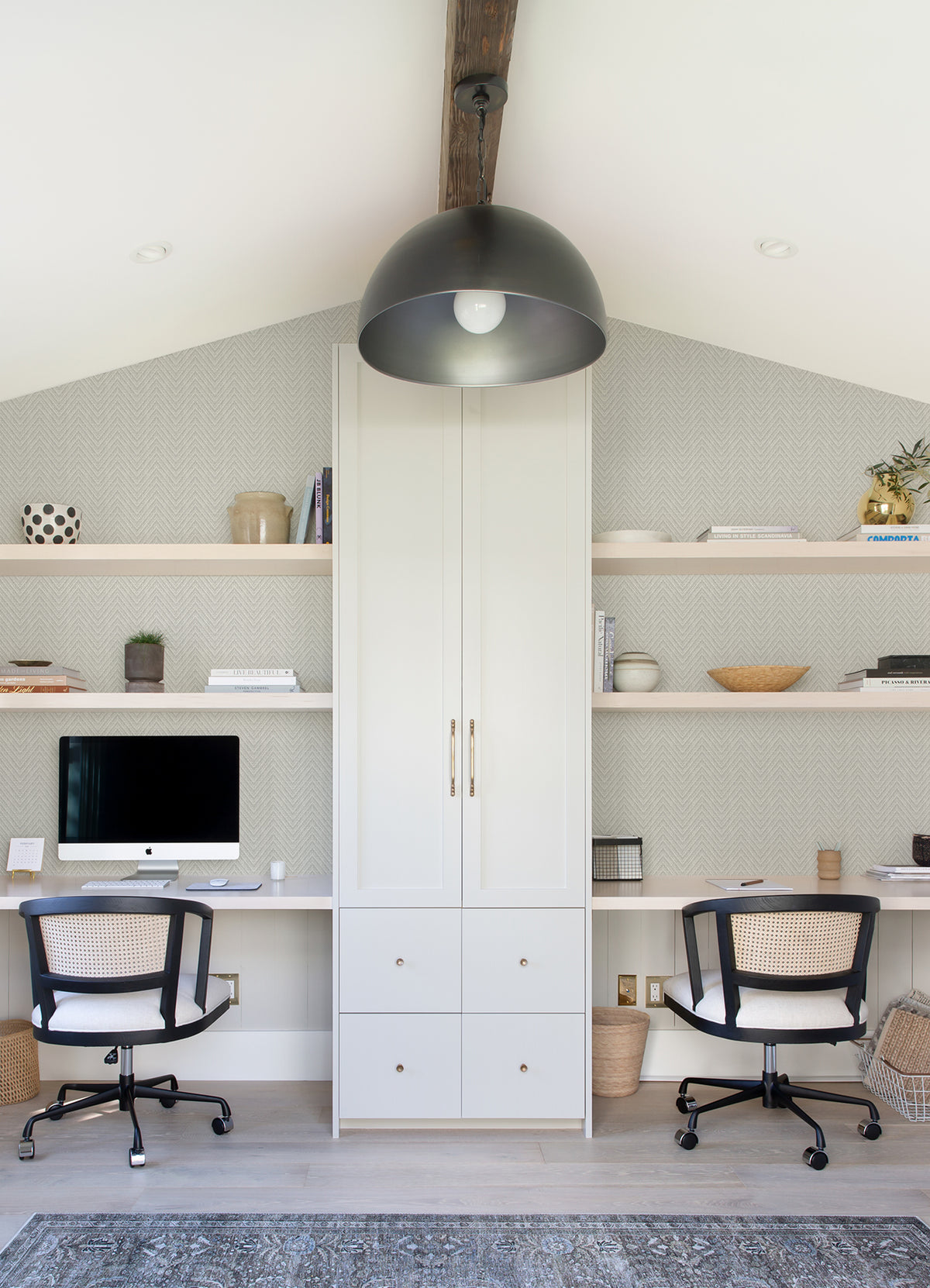 Sure! Here’s a rewritten sentence that incorporates the product data: Modern home office featuring Glynn Chevron Wallpaper by York Wallcoverings, two desks, black chairs, a computer, shelves with décor and books, a central tall cabinet, and a large black light fixture hanging from the ceiling.