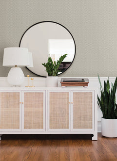 A sideboard with rattan doors sits against a wall adorned with Glynn Chevron Wallpaper by York Wallcoverings. On top of the sideboard are a white lamp, two candlesticks, a potted plant, and a record player. A large round mirror hangs above, while a tall plant in a white pot stands beside the sideboard.