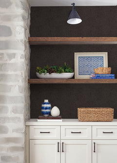 Certainly! Here’s the rewritten sentence incorporating the product data: White cabinets with black handles, floating wooden shelves displaying decor such as a plant, framed art, a woven basket, and blue-and-white vases. A sconce light is mounted above, and the left wall features York Wallcoverings’ Hollenback Dotted Abstract Wallpaper for a stone-look finish.