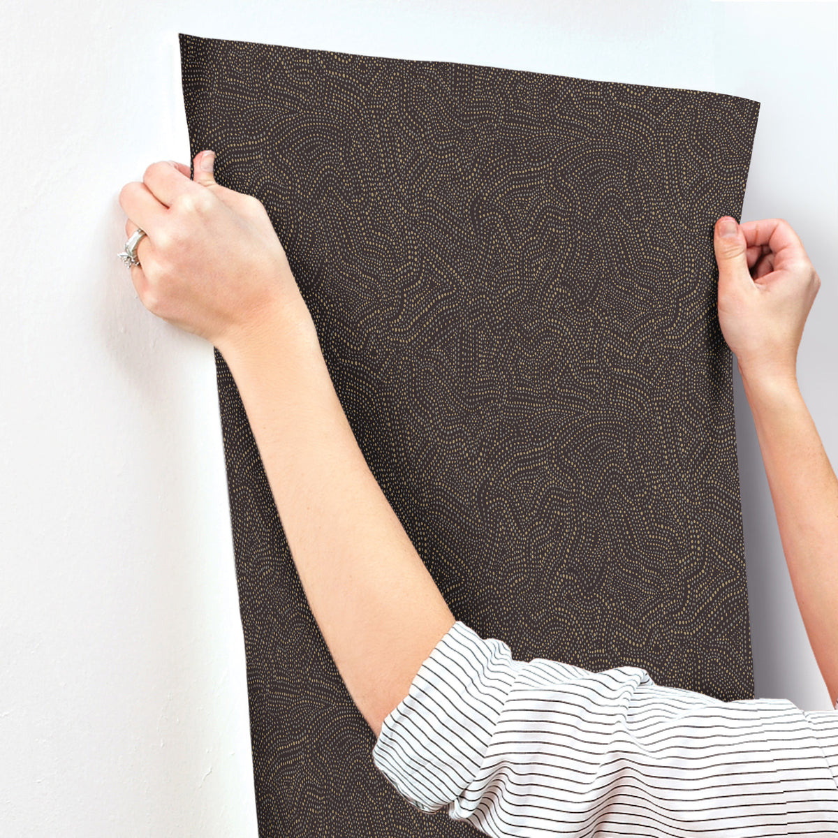 A person in a striped shirt uses both hands to smooth or position the Hollenback Dotted Abstract Wallpaper by York Wallcoverings against a white wall.