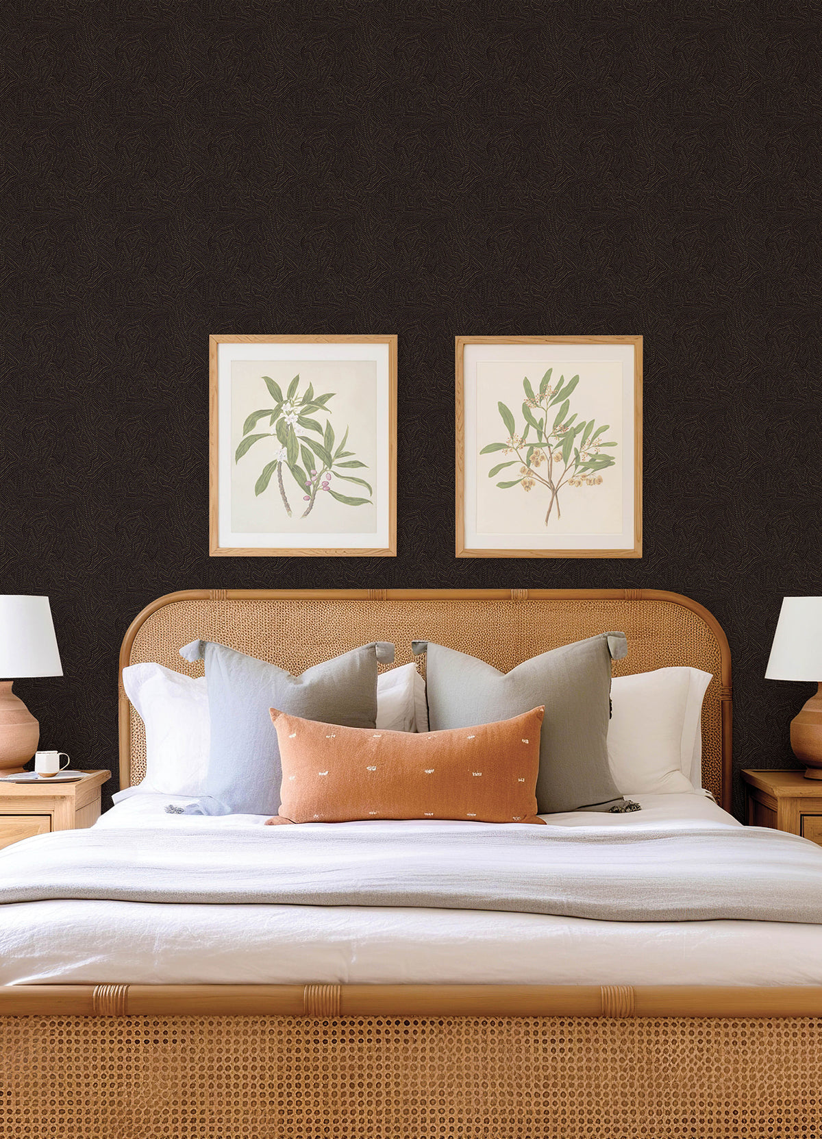 A neatly made bed with neutral pillows and a light brown headboard is centered between two wooden nightstands. Above the bed, two framed botanical prints hang on a dark wall adorned with Hollenback Dotted Abstract Wallpaper by York Wallcoverings.
