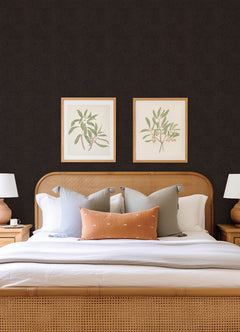 A neatly made bed with neutral pillows and a light brown headboard is centered between two wooden nightstands. Above the bed, two framed botanical prints hang on a dark wall adorned with Hollenback Dotted Abstract Wallpaper by York Wallcoverings.