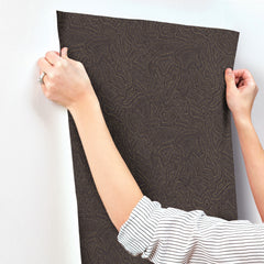A person in a striped shirt holds up York Wallcoverings’ Hollenback Dotted Abstract Wallpaper against a white wall, preparing to apply it.