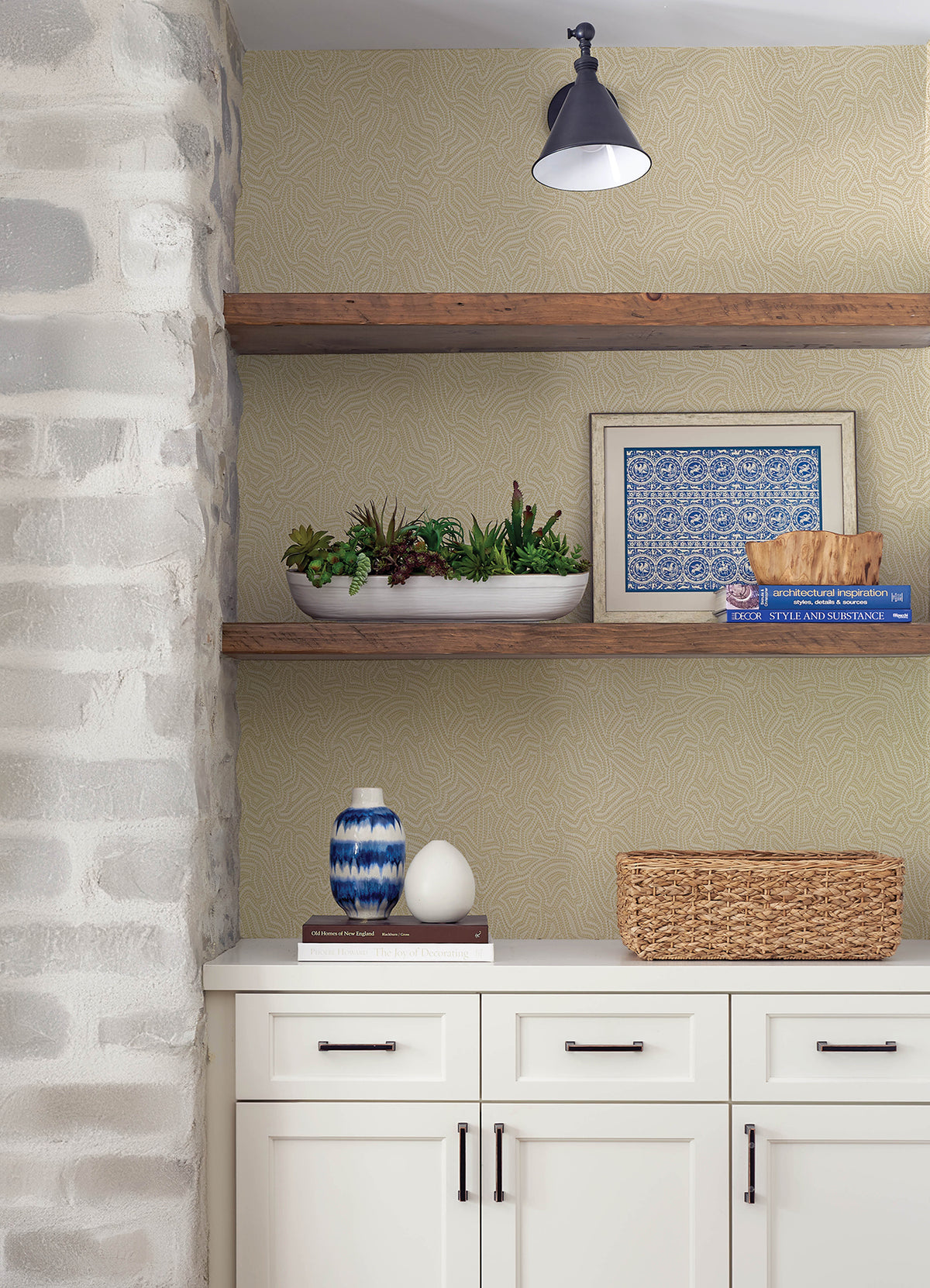 White cabinets with black handles are paired with wooden shelves displaying a framed print, books, a bowl of succulents, vases, and a wicker basket. The left side features Hollenback Dotted Abstract Wallpaper by York Wallcoverings, while above is a small black sconce light.