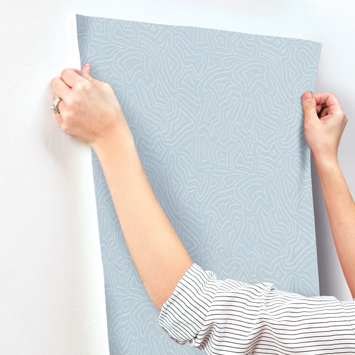 A person in a striped shirt is applying York Wallcoverings’ Hollenback Dotted Abstract Wallpaper, featuring a light blue pattern, to a wall and smoothing it with both hands.