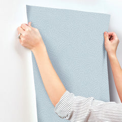 A person in a striped shirt is applying York Wallcoverings’ Hollenback Dotted Abstract Wallpaper, featuring a light blue pattern, to a wall and smoothing it with both hands.