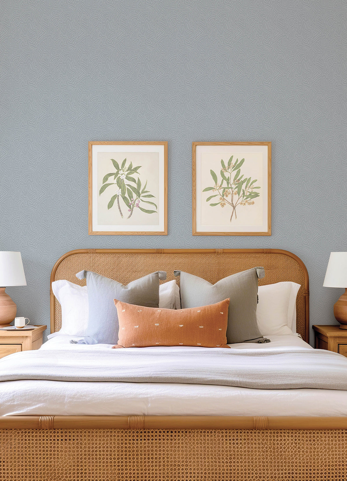 A neatly made bed with neutral pillows and a rust-colored lumbar cushion, flanked by wooden nightstands and lamps, is set against a backdrop of York Wallcoverings’ Hollenback Dotted Abstract Wallpaper in light gray, with two botanical prints hanging above the headboard.