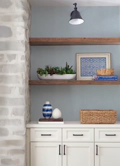 White cabinets, a wicker basket, a decorative blue vase, and books are arranged beneath two wooden shelves displaying a succulent planter, framed art, and a bowl. The scene is set against York Wallcoverings’ Hollenback Dotted Abstract Wallpaper, with part of a stone wall and a black sconce also visible.