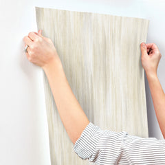 A person wearing a striped shirt applies a sheet of Elysian Ikat Wallpaper by York Wallcoverings—a textured, light-colored wallpaper—to a white wall, smoothing it with their hands. Only their arms and hands are visible.
