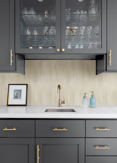 A modern kitchen with dark gray cabinets, gold hardware, a glass-front upper cabinet displaying glassware, a small sink, and soap dispensers atop a white counter. A framed photo leans against a backsplash adorned with York Wallcoverings’ Elysian Ikat Wallpaper.