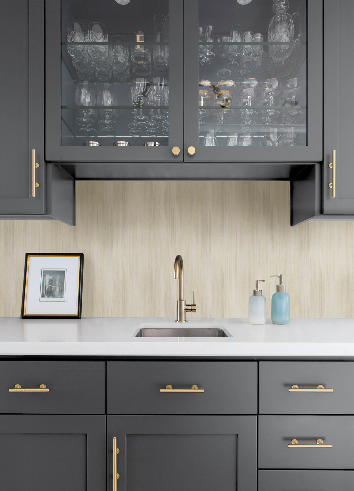 Modern kitchen area with dark gray cabinets, brass hardware, and a white countertop featuring a stainless steel sink. Above the sink, glass-front cabinets showcase glassware, while the walls are adorned with York Wallcoverings’ Elysian Ikat Wallpaper. On the counter, a framed photo and soap dispensers complete the look.