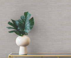 A white vase containing large green monstera leaves sits on a gold console table against neutral-toned Winn Faux Grasscloth Wallpaper by York Wallcoverings.