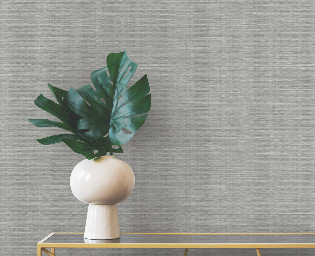 A round white vase containing a large green leaf sits on a gold console table against a gray, horizontally textured Winn Faux Grasscloth Wallpaper by York Wallcoverings.