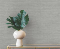 A round white vase containing a large green leaf sits on a gold console table against a gray, horizontally textured Winn Faux Grasscloth Wallpaper by York Wallcoverings.
