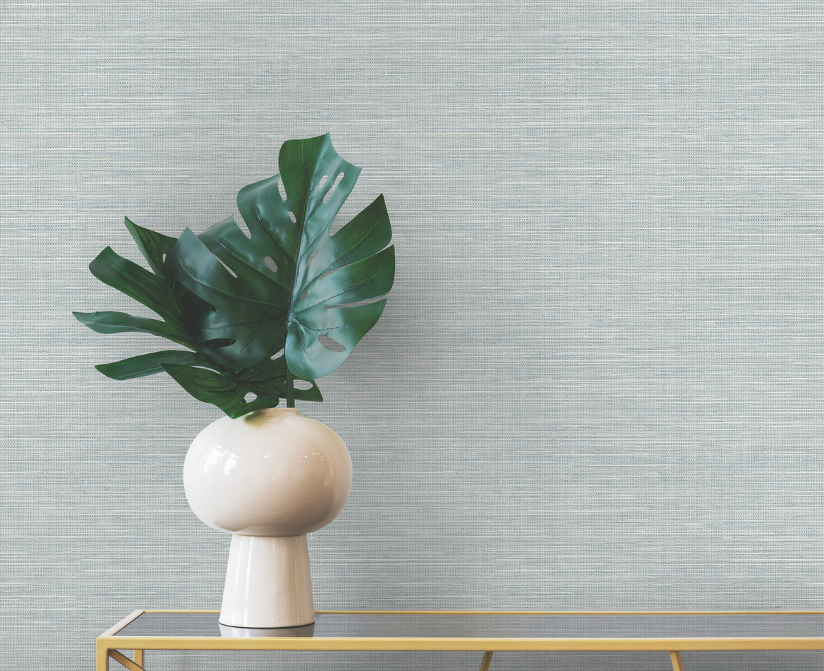 A large monstera leaf in a rounded white vase sits on a gold console table against light blue Winn Faux Grasscloth Wallpaper by York Wallcoverings.