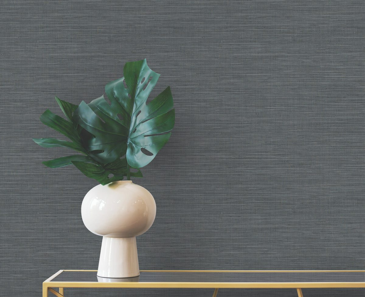 Winn Faux Grasscloth Wallpaper in dark gray provides a backdrop for a white vase containing green monstera leaves on a glass table.