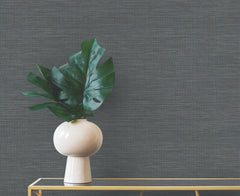 Winn Faux Grasscloth Wallpaper in dark gray provides a backdrop for a white vase containing green monstera leaves on a glass table.