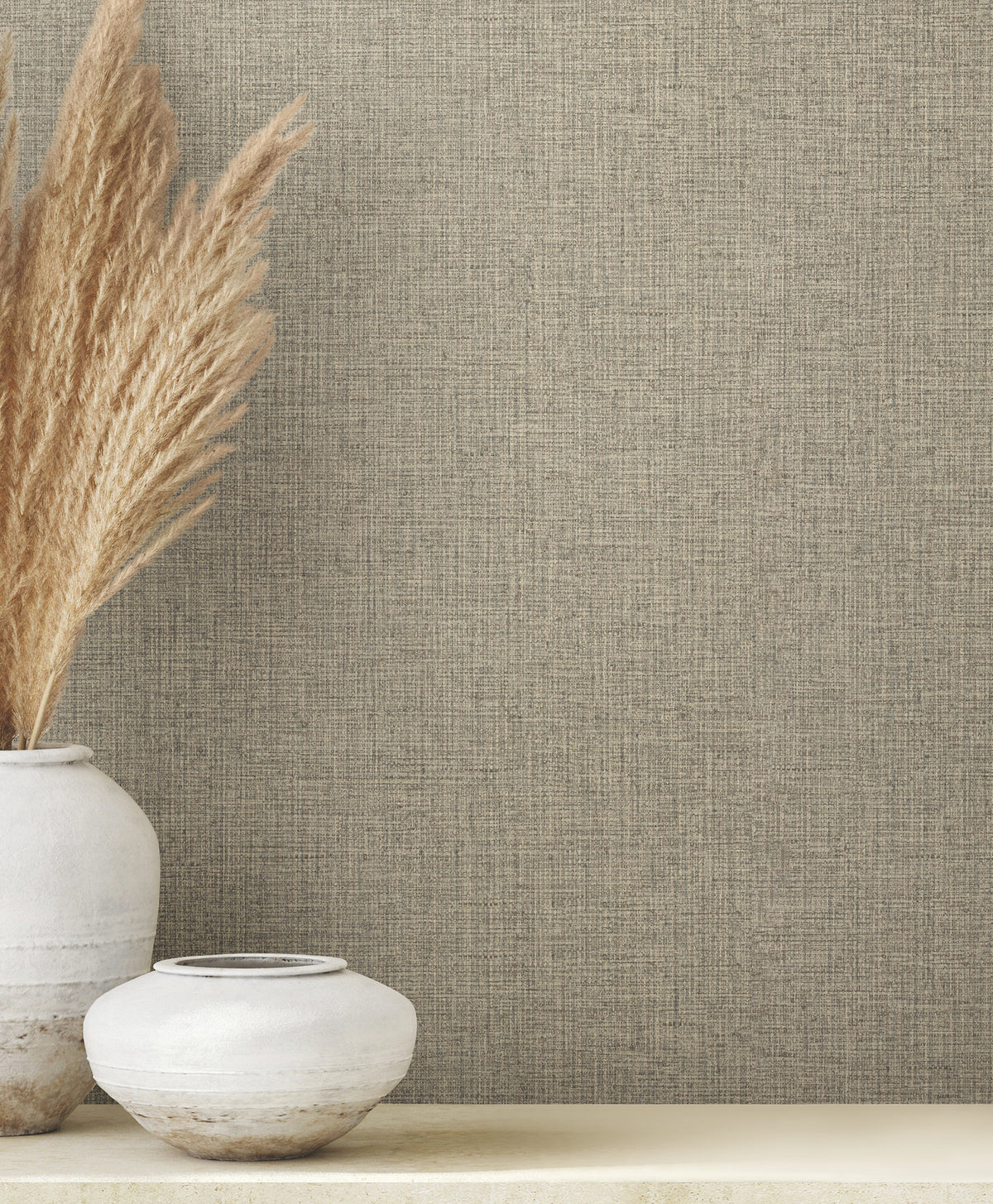 Clarkson Woven Wallpaper in tan with a fine, fabric-like texture shown next to two white ceramic vases, one containing tall dried grass.