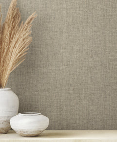 Clarkson Woven Wallpaper in tan with a fine, fabric-like texture shown next to two white ceramic vases, one containing tall dried grass.