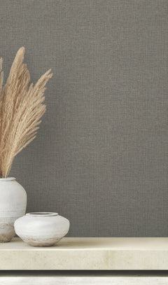 Clarkson Woven Wallpaper in dark gray with a subtle woven texture, shown behind a shelf with two ceramic vases and tall dried grass.