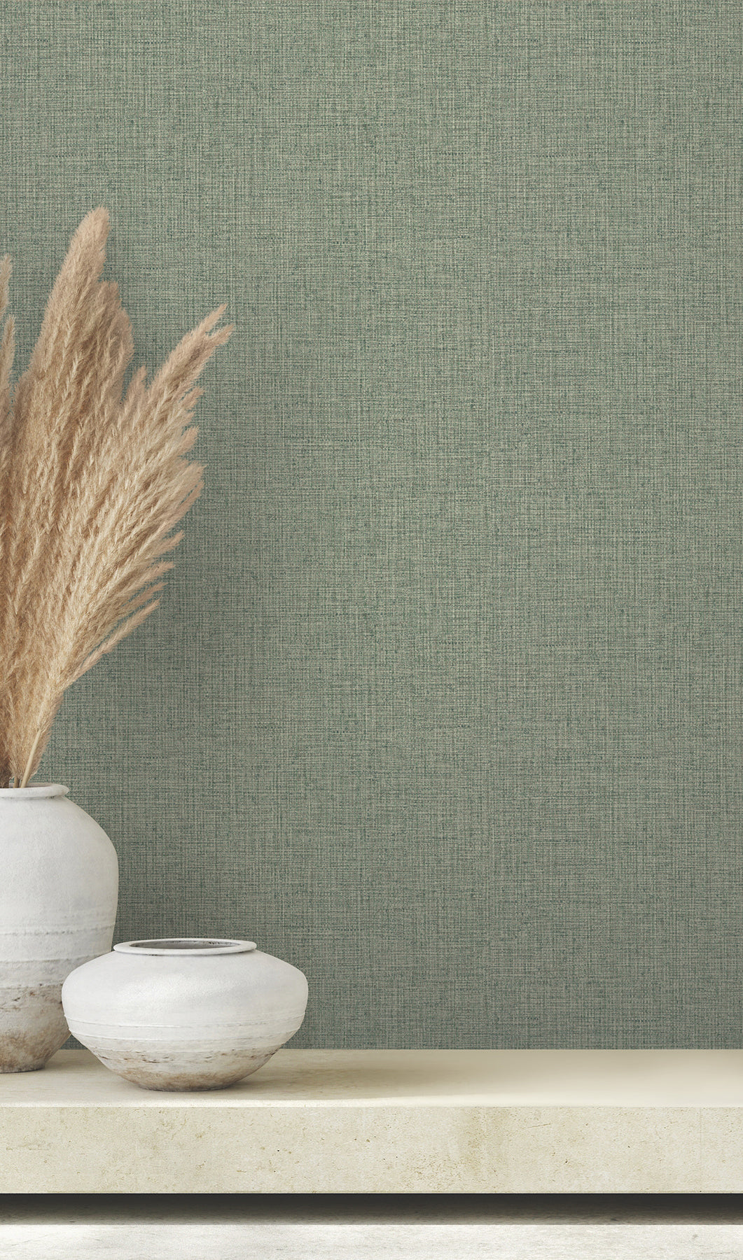 Clarkson Woven Wallpaper in sage green with a woven fabric texture, shown behind a shelf with two ceramic vases and pampas grass.