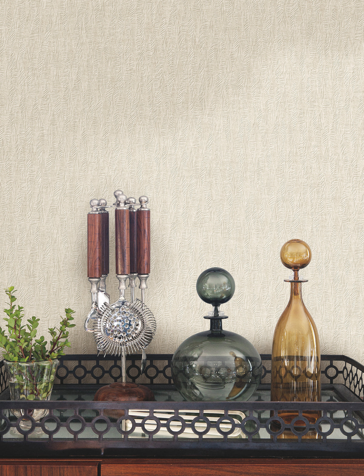 A bar cart with a glass plant vase, a stand of bar tools, and two glass decanters set against light-colored Bryson Feathered Texture Wallpaper by York Wallcoverings with a subtle vertical texture.