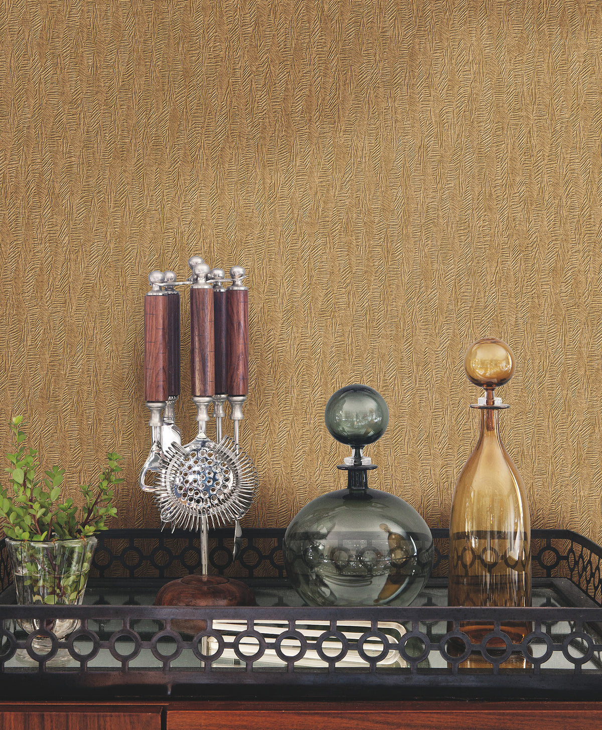Gold-toned Bryson Feathered Texture Wallpaper with a subtle feathered texture behind a bar cart containing a cocktail tool set and two glass decanters.