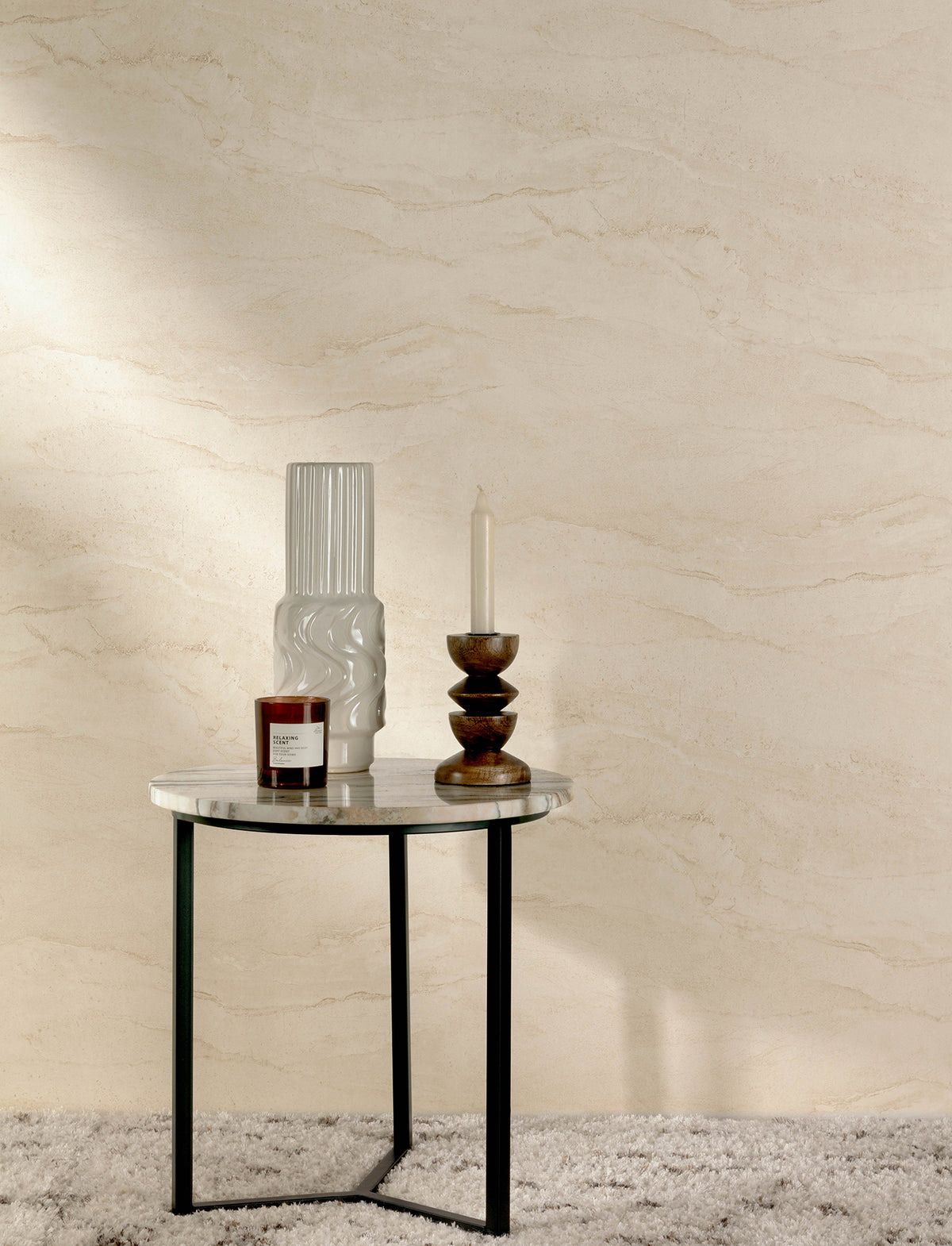 A round marble-top table with black legs holds a white vase, a candle in a glass jar, and a wooden candleholder with a white candle. The table sits on a textured rug against a wall adorned with the Chesapeake Morgana Stone Wallpaper by York Wallcoverings.